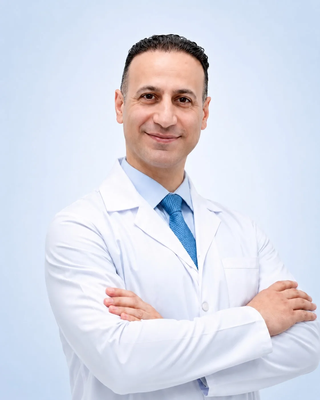 Prof. Dr. Mehmet Emin Şimşek beyaz hekim önlüğü ve mavi kravatıyla, kollarını kavuşturmuş halde açık fonlu stüdyo portresi.