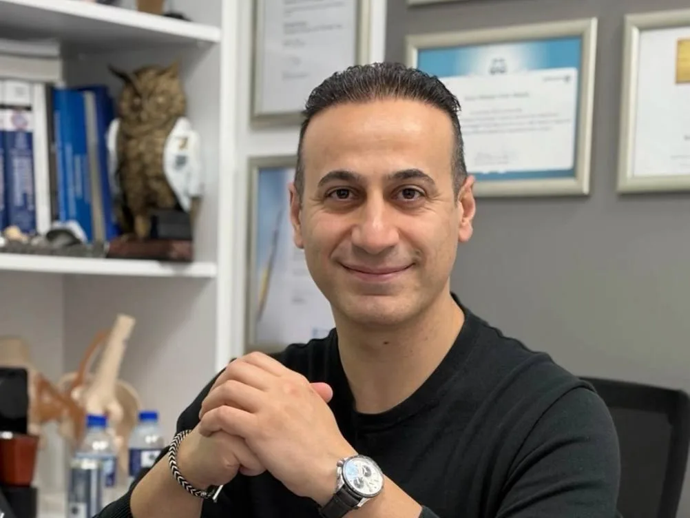 Prof. Dr. Mehmet Emin Şimşek muayenehanesinde, arkasındaki sertifika duvarının önünde çalışma masasında otururken