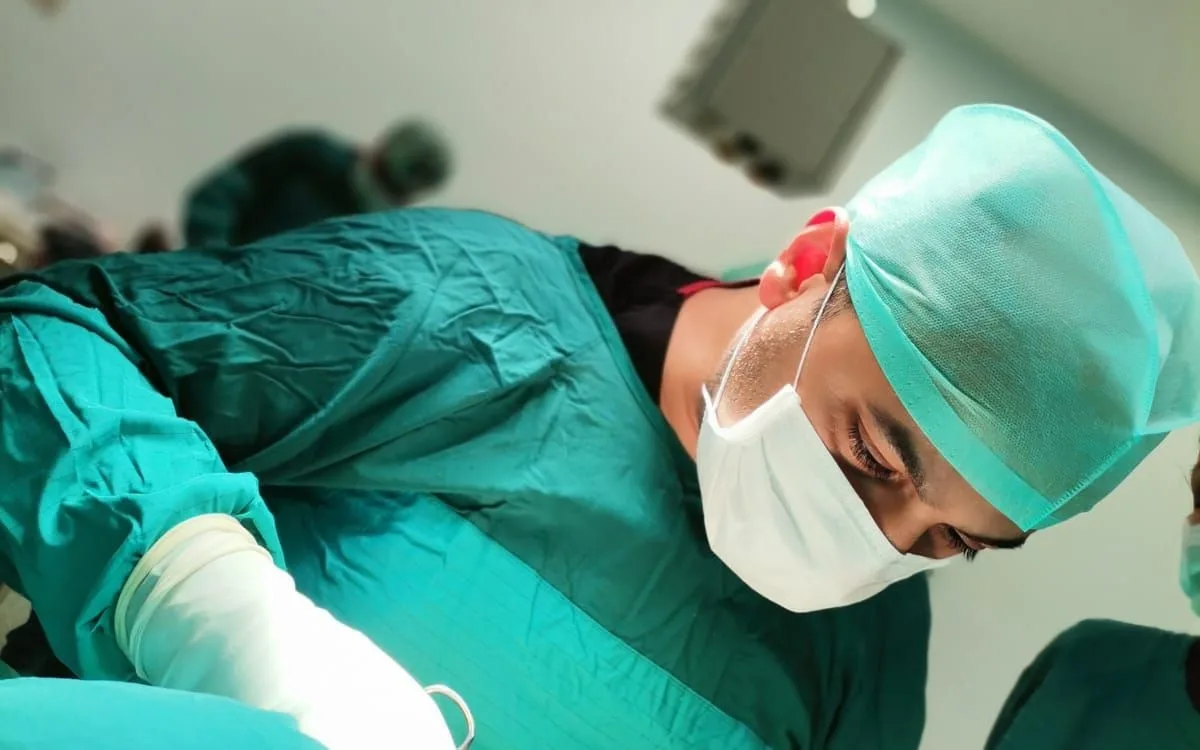 Prof. Dr. Mehmet Emin Şimşek ameliyathanede, bone ve maskeyle cerrahi sahaya eğilmiş.