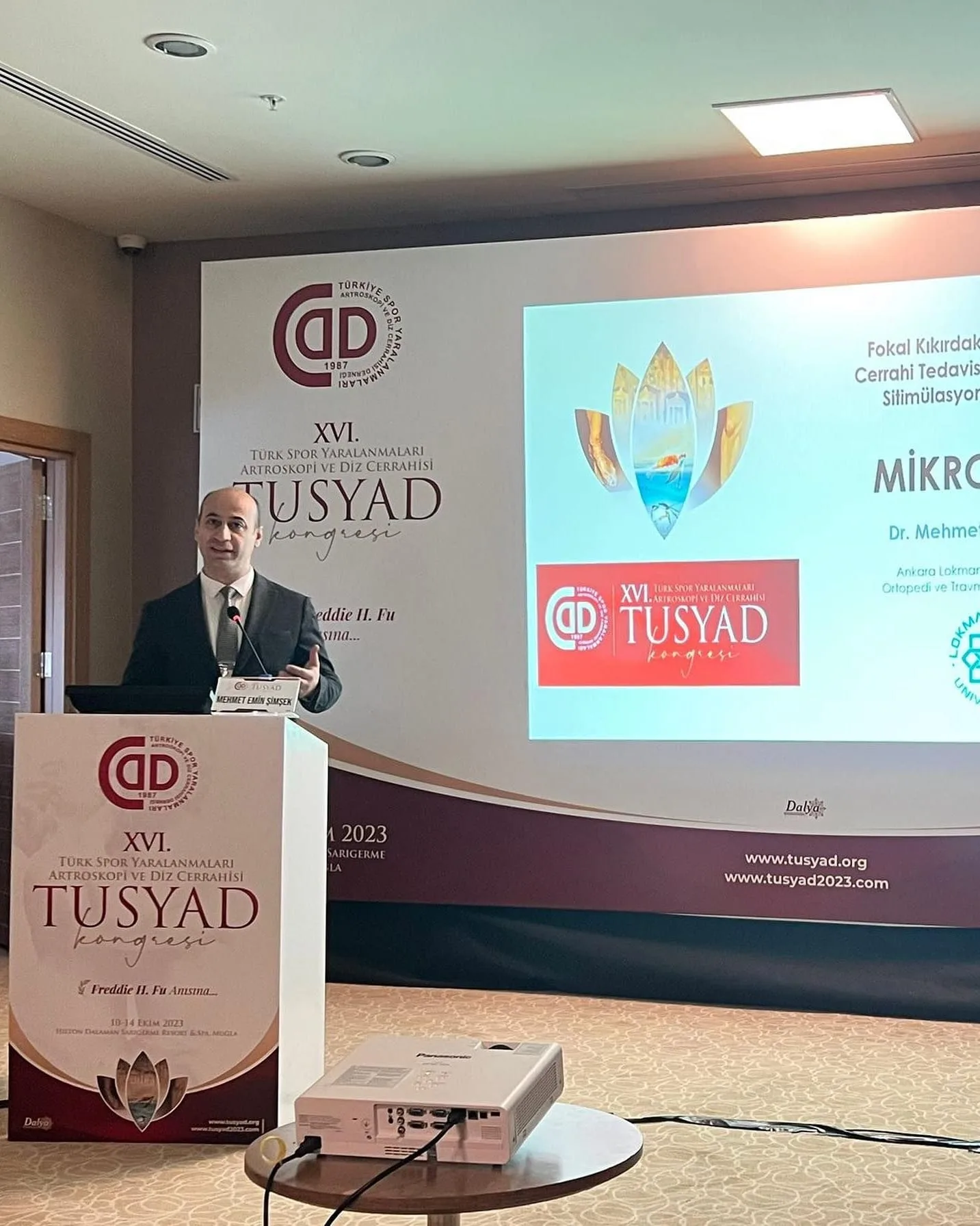 TUSYAD Kongresi kürsüsünde mikrokırık sunumunu yapan hekim ve arkasındaki sunum perdesi