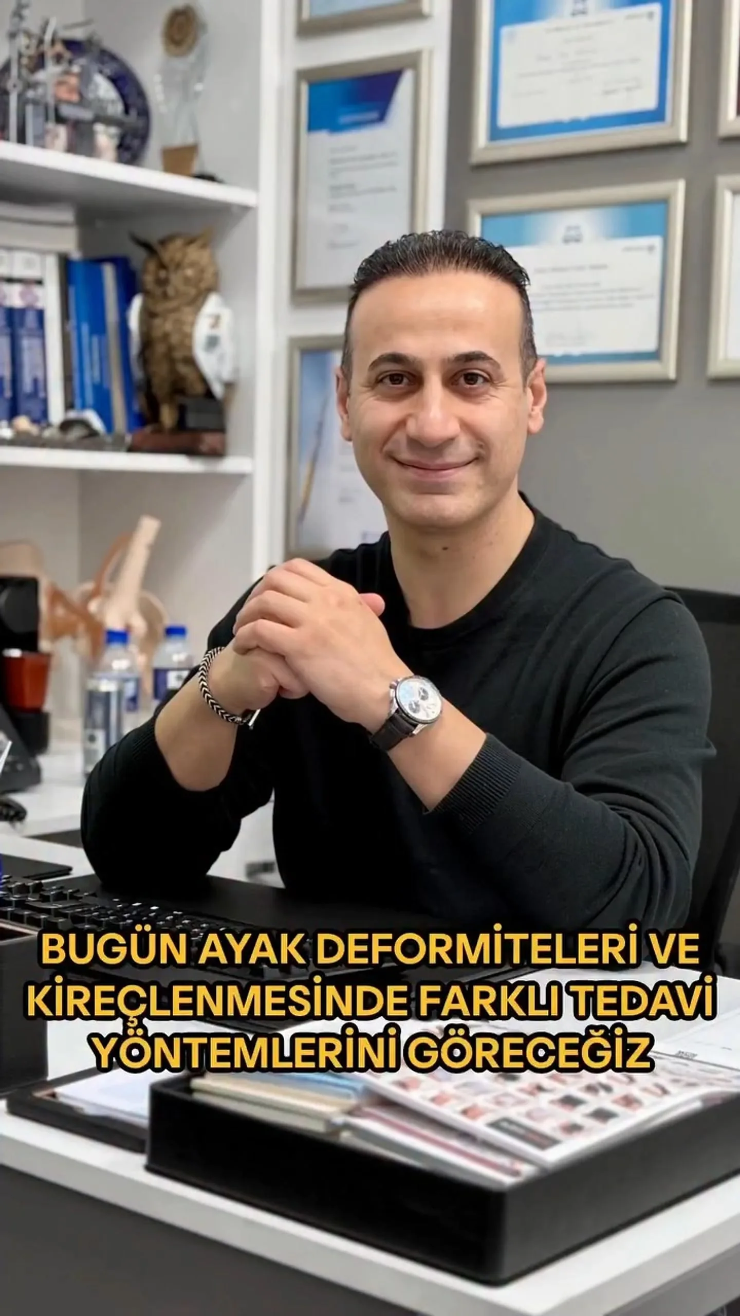 Prof. Dr. Mehmet Emin Şimşek, diploma duvarının önündeki muayenehane masasında otururken